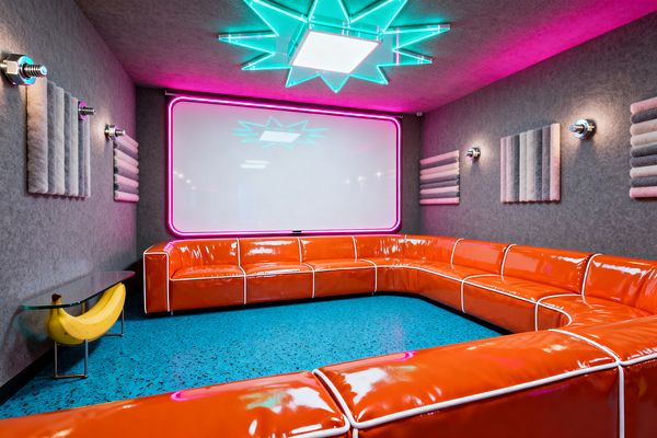 A pop art media room features a glossy tangerine U-shaped sectional with white piping surrounding a blue speckled floor. A large white screen framed in hot-pink neon dominates the front wall beneath a teal acrylic starburst ceiling light. Gray felt walls are lined with ribbed acoustic panels and chrome sconces. At left, a small glass side table sits on a sculptural yellow banana base, adding a playful accent.