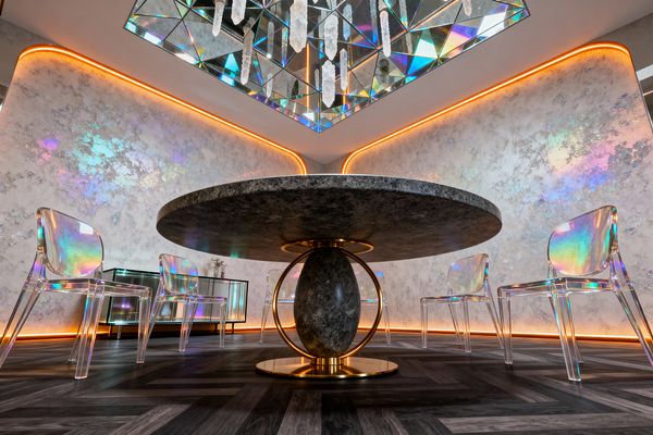 A low-angle view of a futuristic dining room shows a round charcoal stone table with a brass ring base and stone ovoid pedestal, surrounded by four transparent iridescent acrylic chairs. Pearlescent silver walls with rainbow patches and warm orange LED cove lighting frame a faceted mirrored ceiling with hanging crystal pendants; a dark herringbone floor and a low mirrored credenza complete the scene, creating shimmering reflections and a cosmic atmosphere.