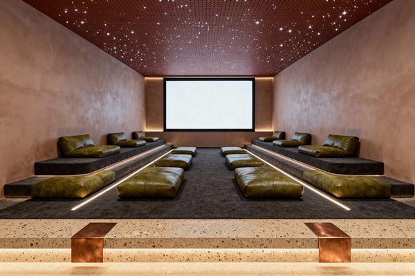 A modern media room with tiered platforms covered in dark charcoal carpet features olive green leather lounge cushions along both sides and low floor pads down the center. Terracotta plaster walls frame a large black-trimmed screen with subtle cove lighting, while a rust-toned perforated ceiling dotted with star-like fiber-optic lights adds ambience. Terrazzo entry steps with copper details and integrated LED step lights complete the warm, cinematic setting.