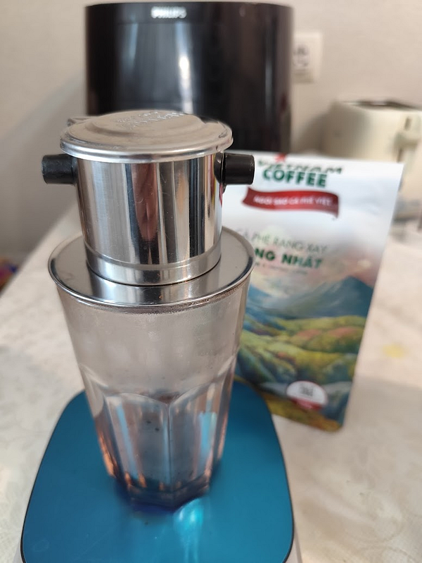 A Vietnamese phin coffee brewer is set up on a blue scale with a glass underneath, alongside a coffee bag.