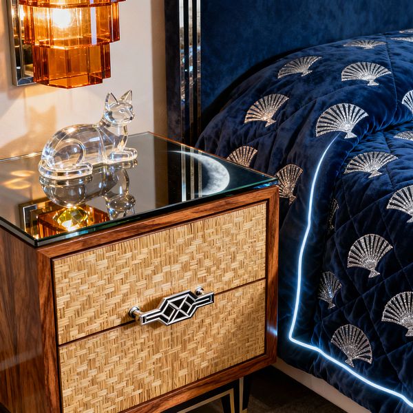 Close-up of an Art Deco bedroom corner with a walnut nightstand featuring woven cane drawer fronts, a mirrored top, chrome geometric pulls, and black-and-gold tapered legs. An amber, tiered glass wall sconce casts warm light onto a clear crystal cat figurine, while a navy velvet quilted coverlet with silver fan motifs and cool LED edge lighting drapes beside a matching velvet headboard framed by polished metal inlays.