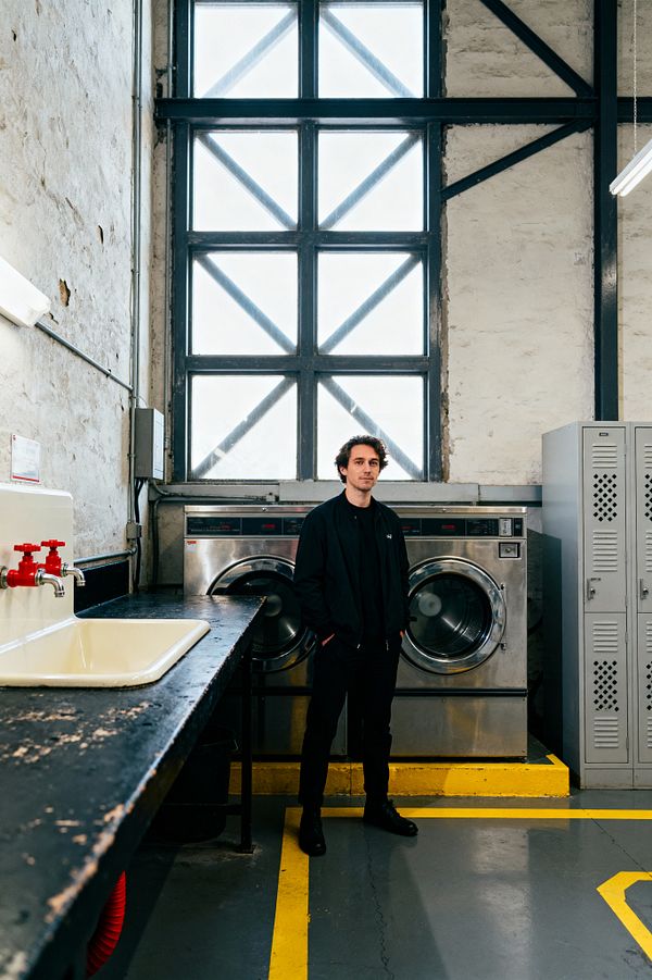 A utilitarian laundry room with industrial finishes: a person stands between two large stainless steel front-loading washers beneath a tall steel-framed window with cross-bracing. To the left, a worn black metal counter holds a cream enamel utility sink with red taps. On the right, light gray perforated metal lockers line the wall. Rough white plaster, exposed conduit, fluorescent lighting, and a gray concrete floor with yellow safety lines define the space.
