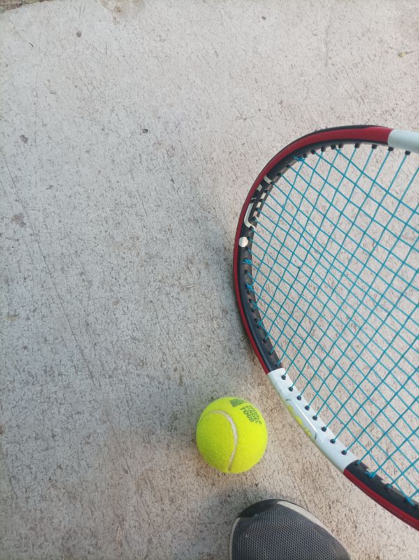 A tennis racket and a bright yellow tennis ball are positioned on a concrete surface.