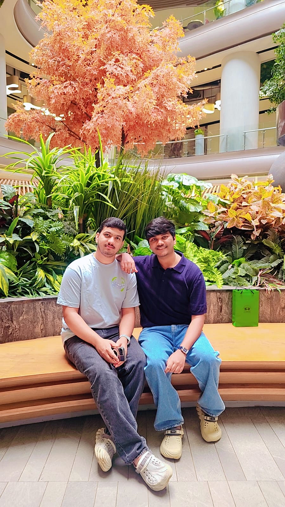 Two young men are sitting on a wooden bench in a vibrant indoor garden area of a mall.