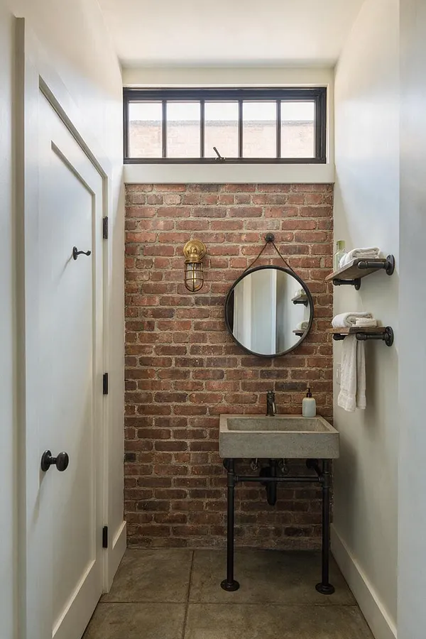 A narrow powder room features a red exposed brick accent wall, a concrete sink on black metal pipe legs, and a round black-framed mirror. White walls, a small transom window, brass sconce lighting, and floating wood shelves with folded towels complete the industrial look.
