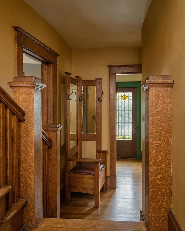 A narrow Craftsman-style entryway with rich wood trim, thick square newel posts, and a built-in bench with dark leather seating. The hall is painted in warm golden tones and leads to a front door with decorative stained glass, creating a cozy, historic feel.