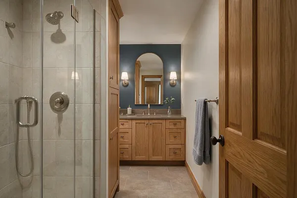 A transitional master bathroom with a glass-enclosed shower on the left, featuring light stone tile, a built-in shower head, and brushed metal fixtures. At the end of the room is a warm oak double vanity with a stone countertop, an arched mirror, and two wall sconces on a deep blue accent wall.