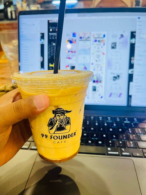 A hand holds a drink from 99 Founder Cafe in front of a laptop displaying various digital designs.