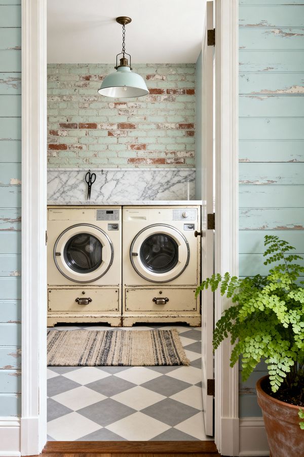 Through a white-trimmed doorway, a compact laundry room shows two cream, front-loading machines with a distressed vintage finish beneath a slim white countertop and a white marble backsplash. A whitewashed mint brick wall and a mint enamel pendant add soft color, while gray-and-white checkerboard tile and a striped rug ground the space; chippy seafoam shiplap frames the view and a terracotta pot with a fern sits by the threshold.