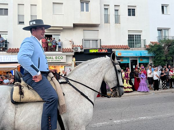 A man in traditional attire rides a white horse during a local festival, with onlookers dressed in colorful outfits in the background.