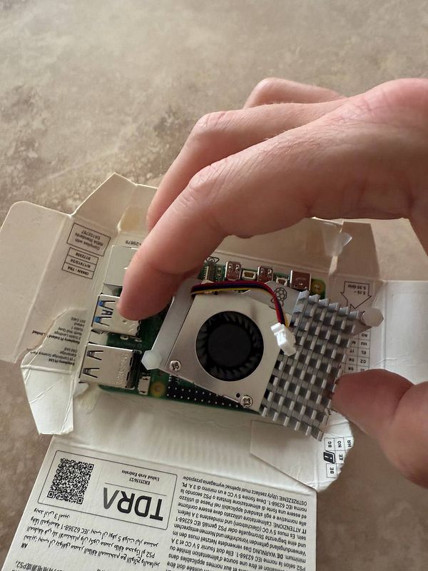 A hand is installing a heatsink onto a Raspberry Pi inside its packaging.