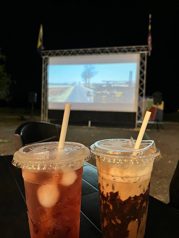 Two drinks are placed on a table in front of a large outdoor screen displaying a scene.