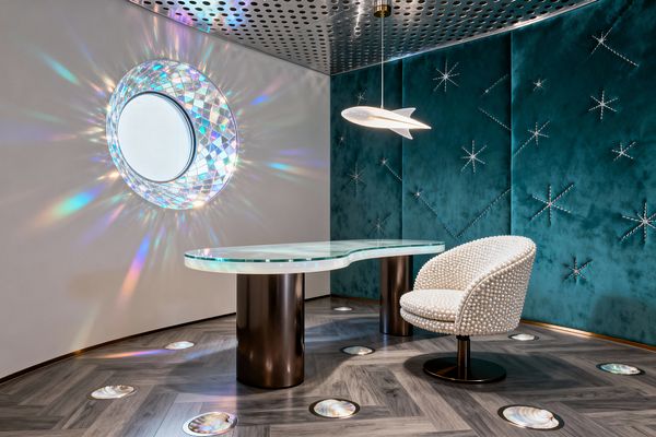Curved home office with a teal velvet feature wall stitched with starburst constellations, a sculptural kidney-shaped glass desk on bronze cylinder legs, and a rounded chair upholstered in pearl-like beads. A circular prismatic window casts rainbow rays across a warm white wall and gray herringbone floor dotted with iridescent round insets. Overhead, a perforated metal ceiling suspends a white rocket-shaped pendant, creating a luminous, space-inspired atmosphere.