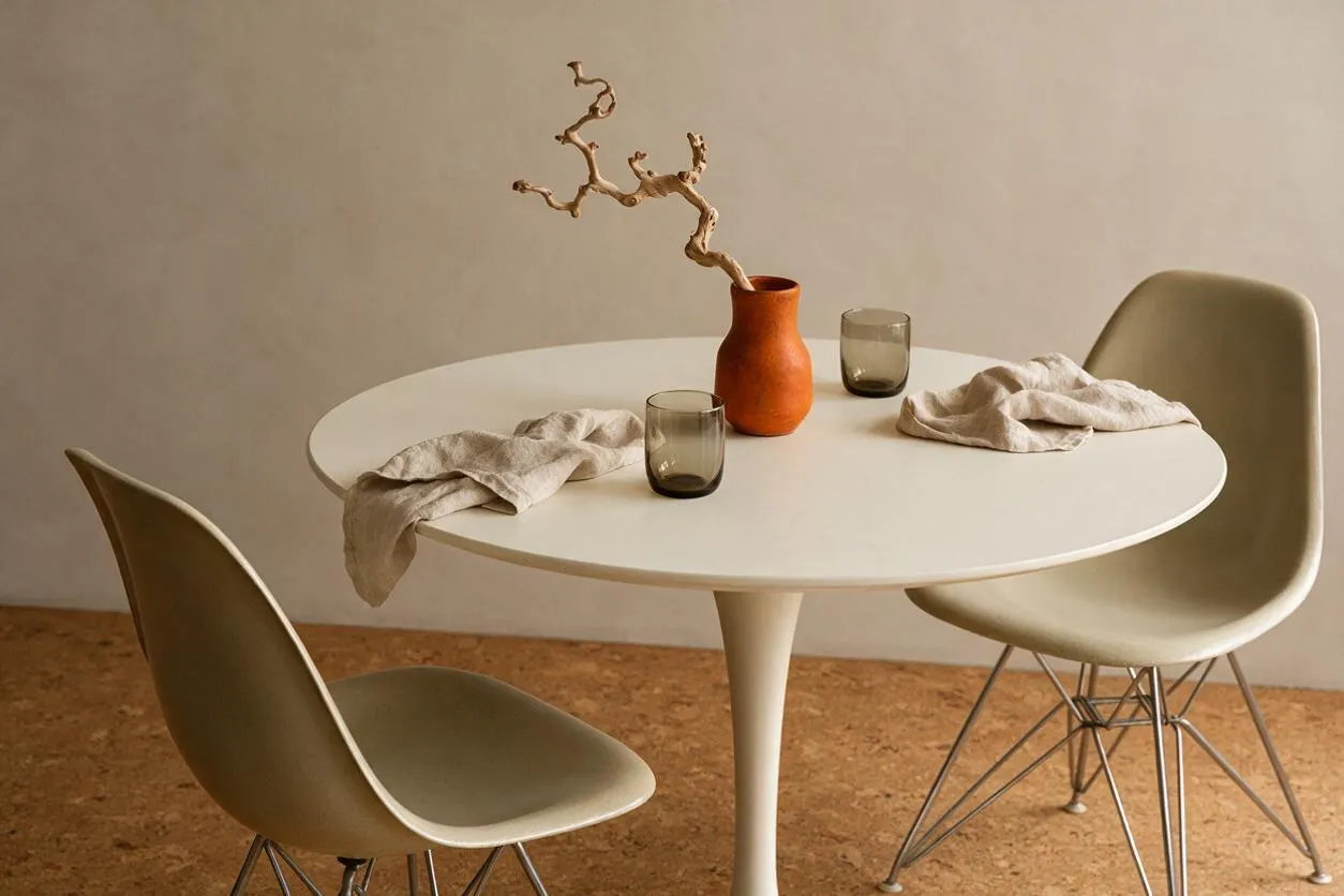 A minimalist dining nook with a round white pedestal table, two molded chairs with wire base legs, and a terracotta vase holding a sculptural branch. Neutral linen drapes across the tabletop beside smoky glass tumblers, while warm beige walls and a cork-toned floor create a calm Mid-Century Modern look.