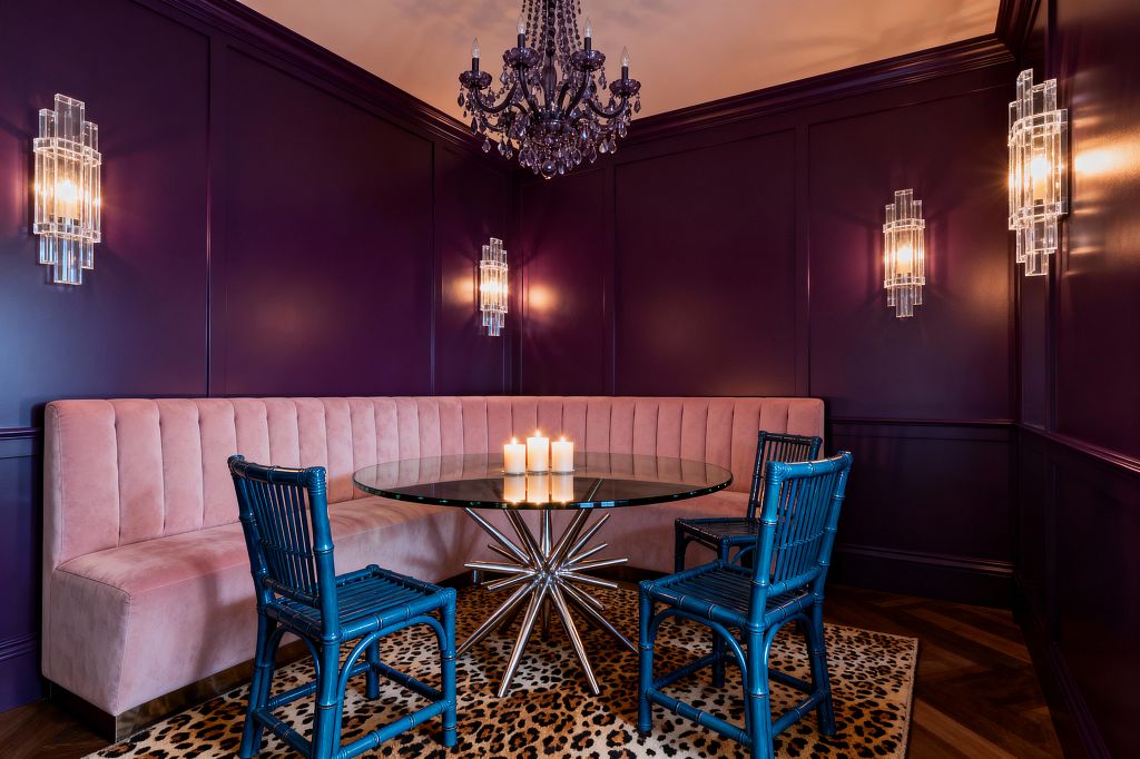 A Hollywood Regency dining nook with deep plum paneled walls features a curved blush velvet channel-tufted banquette surrounding a round glass table with a polished chrome starburst base. Three cobalt blue bamboo-style chairs sit on a leopard-print rug over dark herringbone wood, while crystal Art Deco sconces and a dark glass chandelier cast warm light on a cluster of candles at the table.
