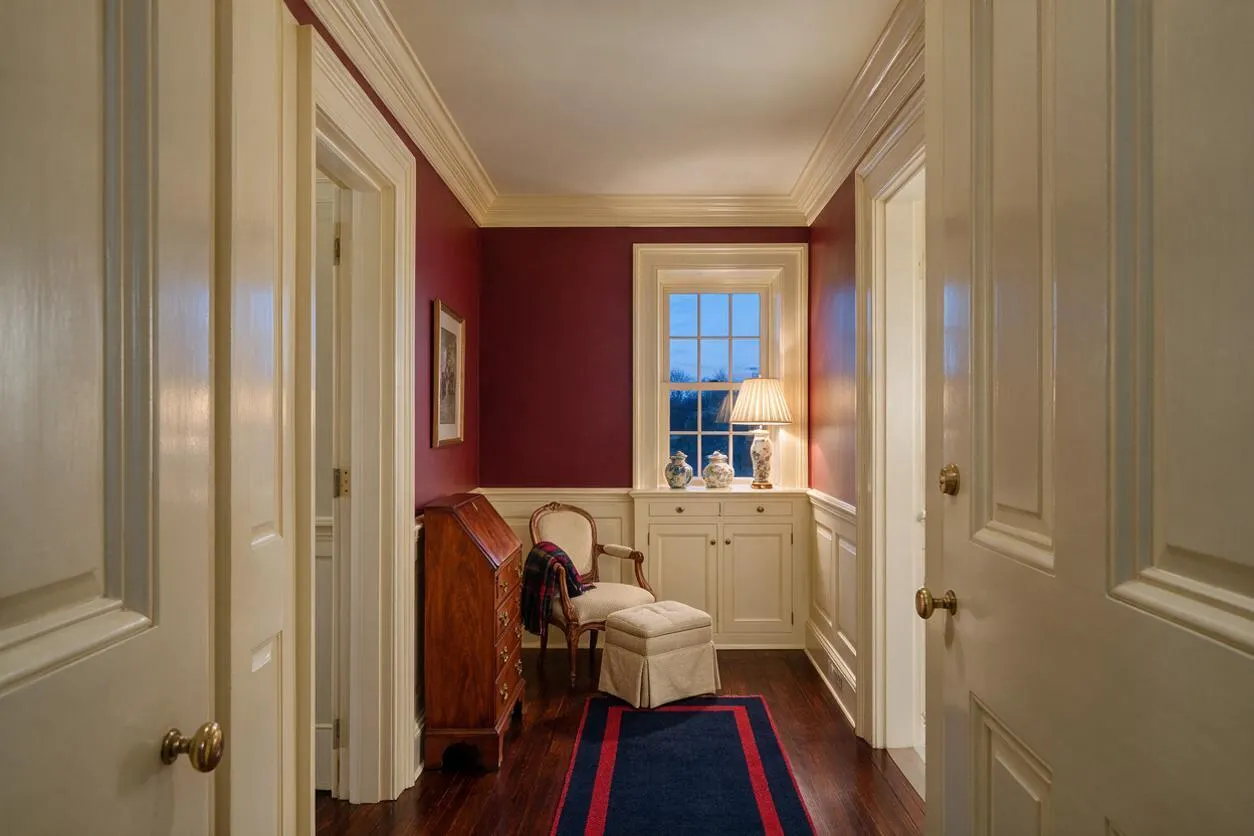 A traditional hallway or front entry space with deep burgundy walls, white crown molding, and paneled wainscoting. A small window with a lamp, upholstered chair, ottoman, and wooden secretary desk create a warm, elegant, lived-in feel.