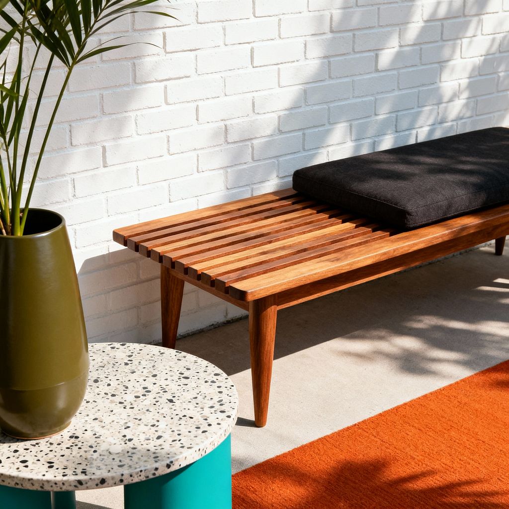 A sunny mid-century modern patio with a white-painted brick wall features a low teak slat bench with tapered legs and a charcoal cushion, set on a smooth concrete floor. In the foreground, a round terrazzo side table with a teal powder-coated base sits beside an olive ceramic planter holding a palm, while a burnt-orange outdoor rug and dappled leaf shadows add warmth and crisp, minimalist geometry.