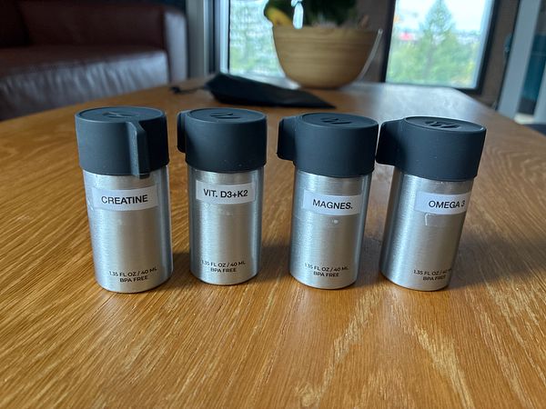 Four small silver supplement containers with black lids labeled with different supplements sit on a wooden table.