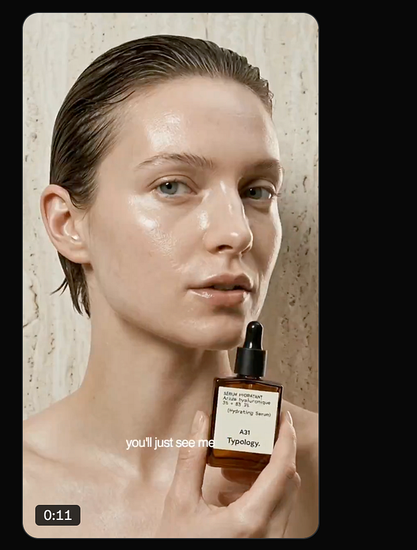 A young woman applies a skincare product to her face in a bright, minimalistic room with large windows in the background.