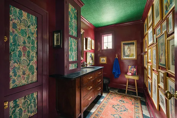A compact powder room with deep burgundy walls, a textured green ceiling, and a gallery wall of framed art covering the right side. The room features a dark wood vanity with a black countertop, patterned fabric or wallpaper panels on the cabinet doors, a small rug, and a blue towel adding a bright accent.
