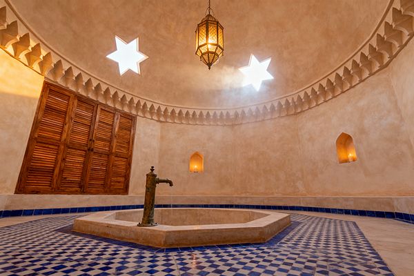 A Moroccan hammam-style bathroom features a domed, sand-colored plaster ceiling punctuated by two star-shaped skylights and a hanging pierced brass lantern. At the center, a sunken stone basin with a patinated brass spout sits on blue-and-white zellige mosaic tiles, while warm tadelakt walls with a sculpted cornice encircle arched candle niches and louvered wooden doors, filling the steamy space with soft, amber light.