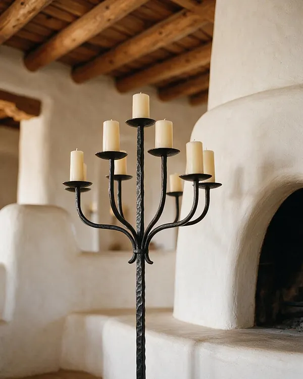 A tall wrought iron candelabra with multiple cream pillar candles stands in front of a smooth white adobe fireplace. The room features thick wooden ceiling beams and soft, earth-toned Southwestern finishes.