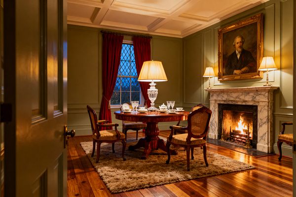 A traditional dining room features sage-green paneled walls, a coffered ceiling, and a round mahogany pedestal table with four cane-back chairs on a plush beige rug. A crystal lamp with a pleated shade sits at the table’s center, while two lamps flank a white marble fireplace with a lit fire and a gilded portrait above. Deep red drapes frame a leaded window, and wide-plank wood floors warm the scene.
