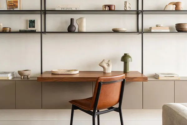 A modern studio apartment workspace with a long walnut floating desk, black metal shelving above, and taupe built-in cabinets below. The shelves are styled with neutral ceramics, books, and decor, while a tan leather chair sits in front, creating a warm, minimal interior.