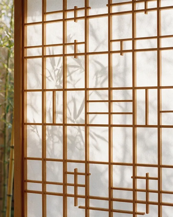 A close-up of a Japanese Zen-style shoji screen with a light wood lattice over translucent paper. Soft bamboo shadows fall across the panels, creating a calm, natural atmosphere with warm daylight filtering through.