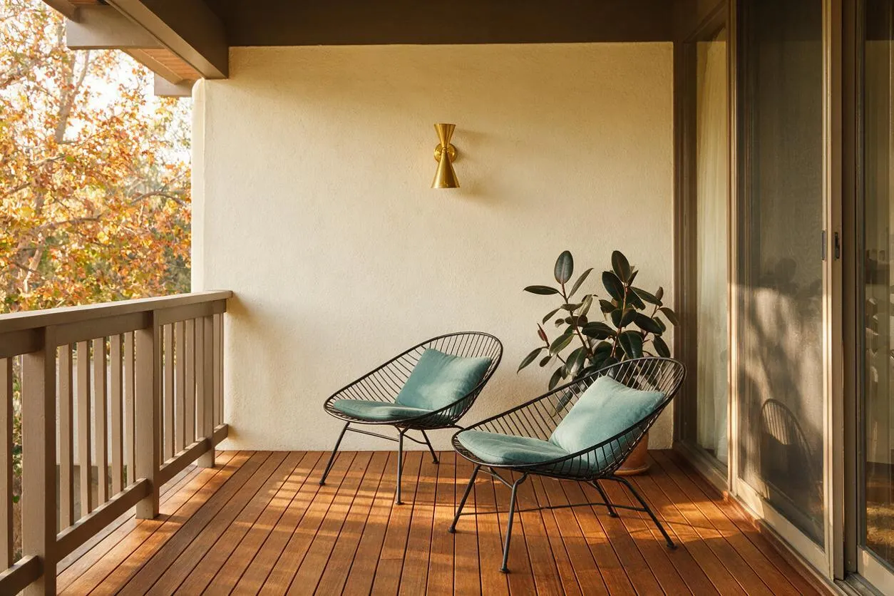A small covered balcony in mid-century modern style with warm wood decking, a simple cream stucco wall, and a brass wall sconce. Two black wire lounge chairs with pale teal cushions sit near a potted rubber plant, while a wood railing and sliding glass door frame the space with soft autumn light.