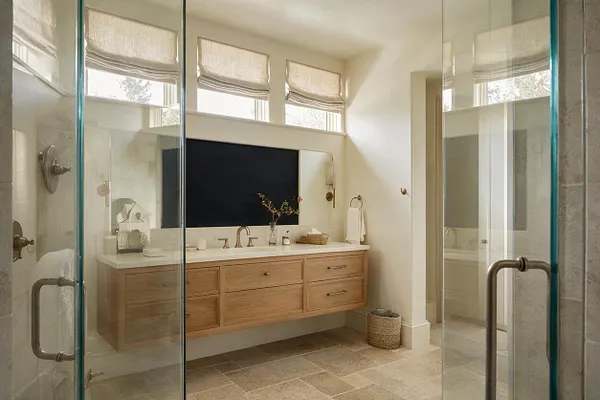 A bright transitional master bathroom with a floating light oak double vanity, a large rectangular mirror, and soft neutral walls. Glass shower doors frame the view, while three high windows with Roman shades bring in natural light over the stone tile floor.