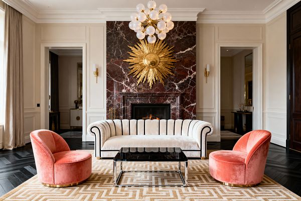 A Hollywood Regency living room centers on a burgundy marble fireplace with a gilded sunburst above and a brass globe chandelier overhead. An ivory velvet, scalloped sofa with black piping faces a chrome and black-glass coffee table, flanked by two coral velvet barrel chairs on brass plinth bases. Cream paneled walls, brass sconces, a beige Greek key rug, and dark herringbone wood floors create a symmetrical, glamorous setting around a lit fire.