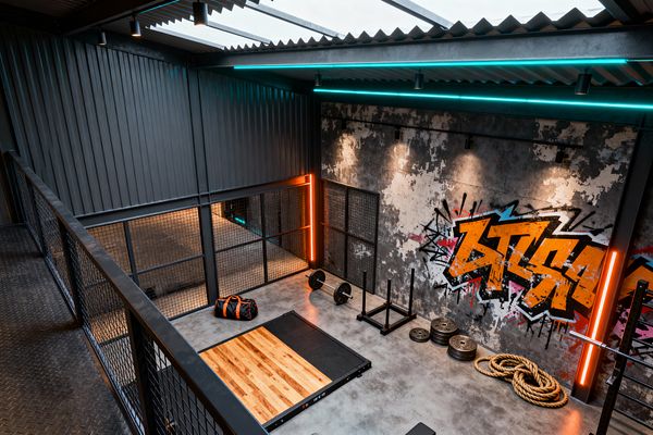 View from an upper catwalk shows a compact home gym wrapped in corrugated steel with exposed beams and skylight roof panels. A hardwood lifting platform set in black rubber anchors the concrete floor, with stacked weight plates, a sled, battle ropes, and a loaded barbell nearby; a distressed wall with bold orange and teal graffiti is illuminated by track lights, cyan strips, and vertical orange LED bars.