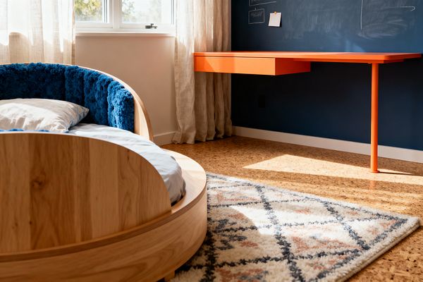 A contemporary kids’ room features a curved light-oak bed with deep blue textured upholstery and pale gray bedding near a sunlit window with sheer linen curtains. A bright orange, wall-mounted desk with a single support leg spans a navy chalkboard wall, while warm cork flooring and a cream diamond-patterned rug in slate and terracotta tones create a playful, tidy workspace and sleep nook.
