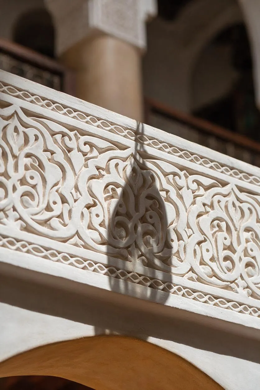 A close-up of an ornate white carved railing with swirling floral arabesque patterns and delicate lattice borders. Soft shadows fall across the textured surface, with blurred arches and a warm beige wall in the background.