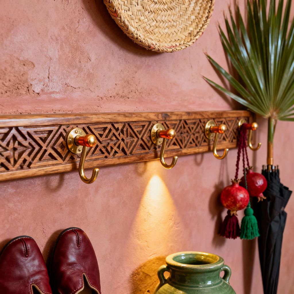 A Moroccan-inspired mudroom vignette with a rosy terracotta plaster wall and a carved wooden rail fitted with five brass hooks, each capped with small amber-toned knobs. A spotlight washes the wall below. Woven straw hat, palm frond, and pomegranate tassels hang nearby, alongside a black umbrella. At the bottom, burgundy leather slippers rest beside a glossy green ceramic jar. The scene feels warm and artisanal, with layered textures and geometric motifs.