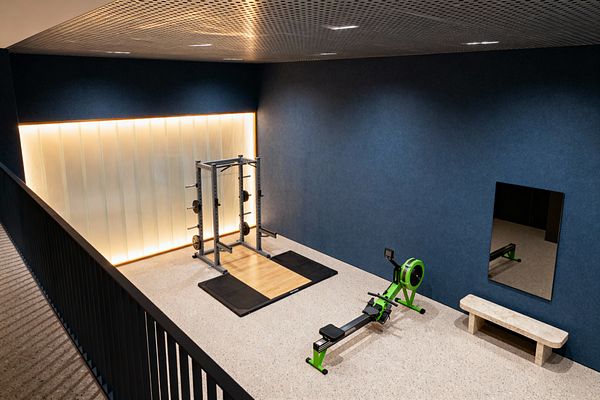 Contemporary home gym with a steel power rack set on a wood-and-black rubber lifting platform and a bright green rowing machine on speckled terrazzo flooring. A deep navy wall holds a rectangular mirror and stone bench, while a ribbed translucent backlit panel and perforated metal ceiling with recessed lights create warm, even illumination beside a slim black balustrade.