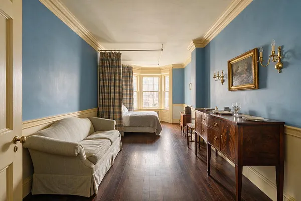 A narrow traditional studio apartment with blue walls, cream trim, and dark wood floors. The space includes a beige sofa on the left, a bed near a bay window with plaid curtains, and a mahogany sideboard with brass accents and wall sconces on the right.