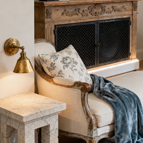 A close-up of a French Country media room corner featuring an ivory linen chaise with a distressed, carved wood frame, accented by a gray floral pillow and a slate-blue velvet throw. A square honed marble side table stands beside a patinated brass wall sconce on a warm white wall. In the background, a mantel-style carved wood media cabinet with black iron mesh doors hides speakers, softly underlit by LED strip lighting over a wood floor.