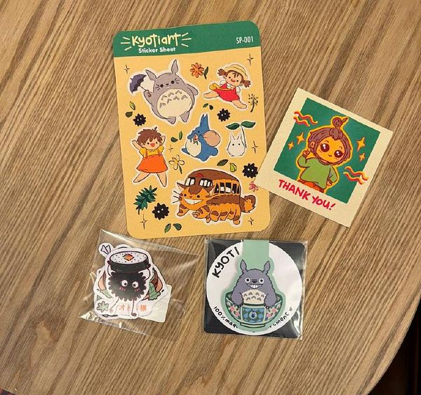 A collection of colorful stickers and a thank you card from kyoti.art displayed on a wooden surface.