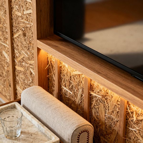 Close-up of a Scandinavian media room featuring a flat-screen TV recessed into an oak wood frame. Behind the screen, textured wood-wool acoustic panels sit between vertical slats and are washed by warm LED strip lighting. In the foreground, a beige linen bolster with contrast stitching rests beside a pale marble tray holding a clear tumbler. The palette is warm and neutral, emphasizing natural textures and clean, crafted details.