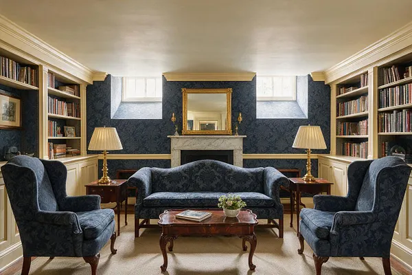 A traditional basement sitting room with dark blue patterned wallpaper, built-in bookshelves on both sides, and a centered marble fireplace with a gold-framed mirror above it. The room includes a blue sofa, two matching wingback chairs, wood side tables with brass lamps, a carved coffee table, and small basement windows near the ceiling.
