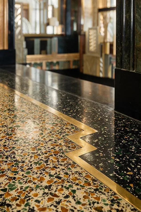 A close-up view of an Art Deco-inspired floor shows a speckled terrazzo surface in cream, amber, green, and black tones divided by a zigzag brass inlay. A glossy black terrazzo band runs beside it, while blurred mirrored and metallic wall details create a luxurious backdrop.