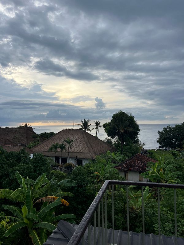 A scenic view of a coastal area in Bali, featuring traditional thatched-roof buildings and lush greenery under a cloudy sky.