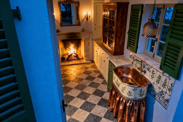 A cozy French Country kitchen features a lit fireplace with a carved stone mantel and a gilded mirror above. Cream cabinets and a glass-front wood hutch run along one wall. A copper farmhouse sink with a floral ceramic apron and a brass wall-mounted faucet sits beneath windows with green shutters and a perforated pendant light. Light stone counters and a beige-and-gray checkerboard stone floor complete the warm, rustic setting.