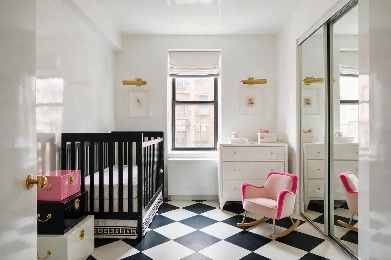 A bright nursery with glossy white walls, a black crib with decorative trim, and a black-and-white checkered floor. Pink and white storage trunks, a blush rocking chair with hot pink trim, mirrored closet doors, and gold accents add a glamorous Hollywood Regency feel.