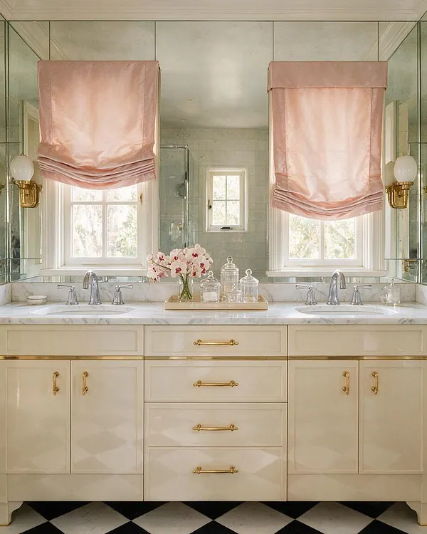 A Hollywood Regency master bathroom with a large double vanity in creamy lacquered cabinetry, marble countertops, and polished brass hardware. Two windows are framed by blush pink Roman shades, with mirrored walls, twin sinks, and black-and-white checkered flooring adding glamour and symmetry.