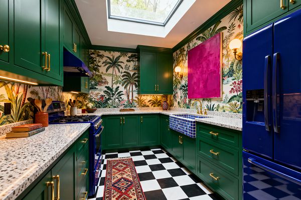 U-shaped kitchen with emerald green Shaker cabinets and terrazzo counters, wrapped in tropical palm wallpaper. A cobalt blue range and refrigerator sit opposite a blue grid-tiled apron-front sink with a brass bridge faucet, beneath a large skylight; a magenta abstract painting and globe sconces add warmth, while a black-and-white checkerboard floor and a patterned runner ground the space.