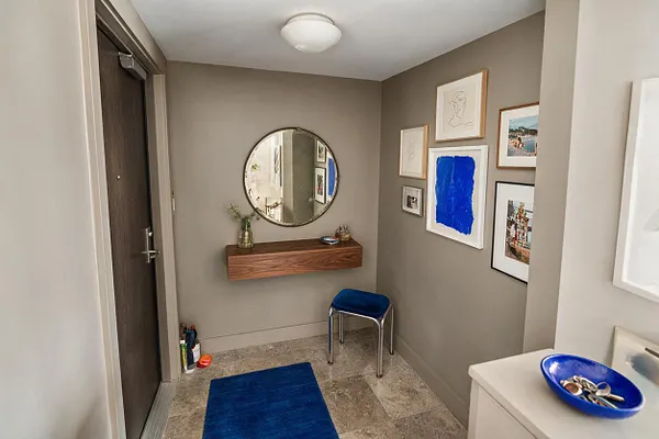 A small eclectic entryway with taupe walls, a dark wood front door, and a round wall mirror above a floating wood console shelf. The space includes a bright blue stool, a blue runner rug, and a gallery wall of mixed framed art, creating a layered, collected look.