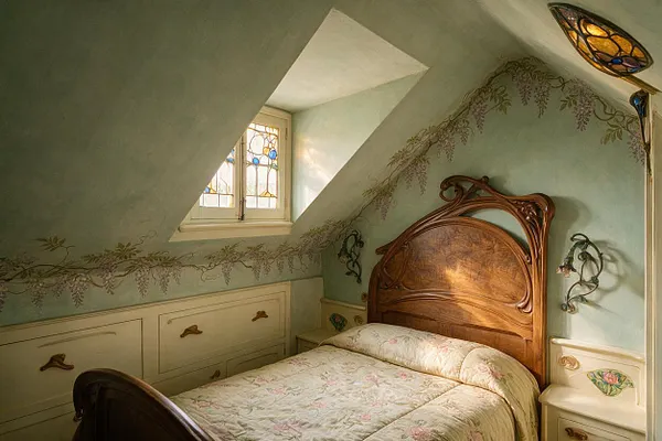 A cozy attic bedroom in Art Nouveau style with a carved wooden bed, floral quilt, and pale mint walls decorated with vine-like mural details. A small stained-glass window and soft built-in cabinetry complete the intimate, decorative space.